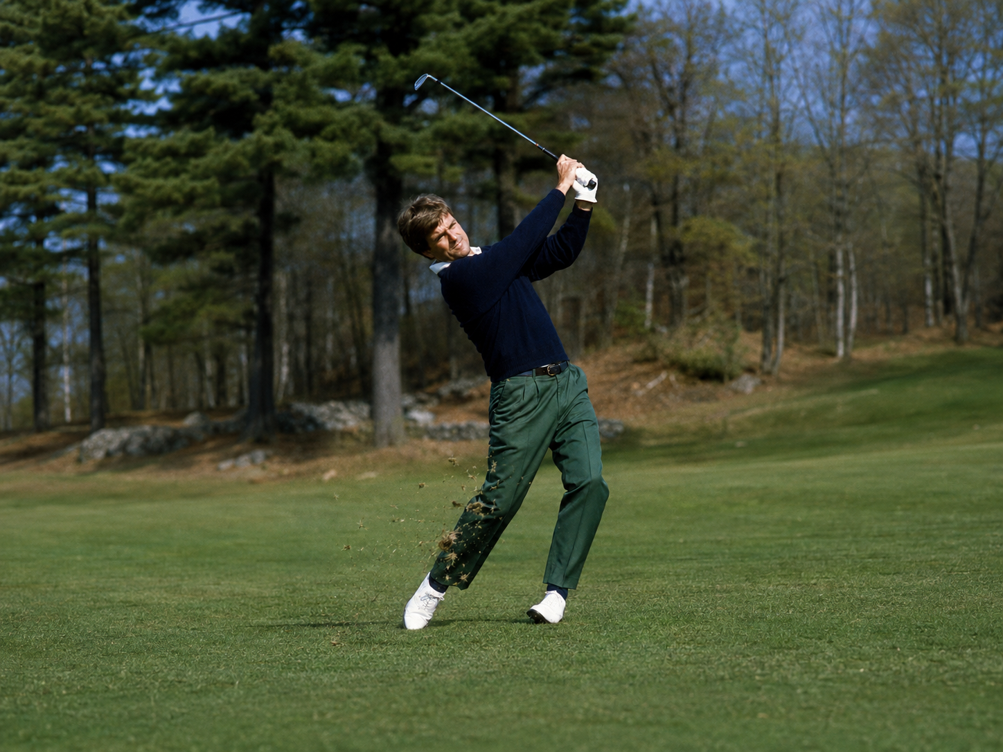 A member finishing an iron shot from the fairway, tree line beyond.